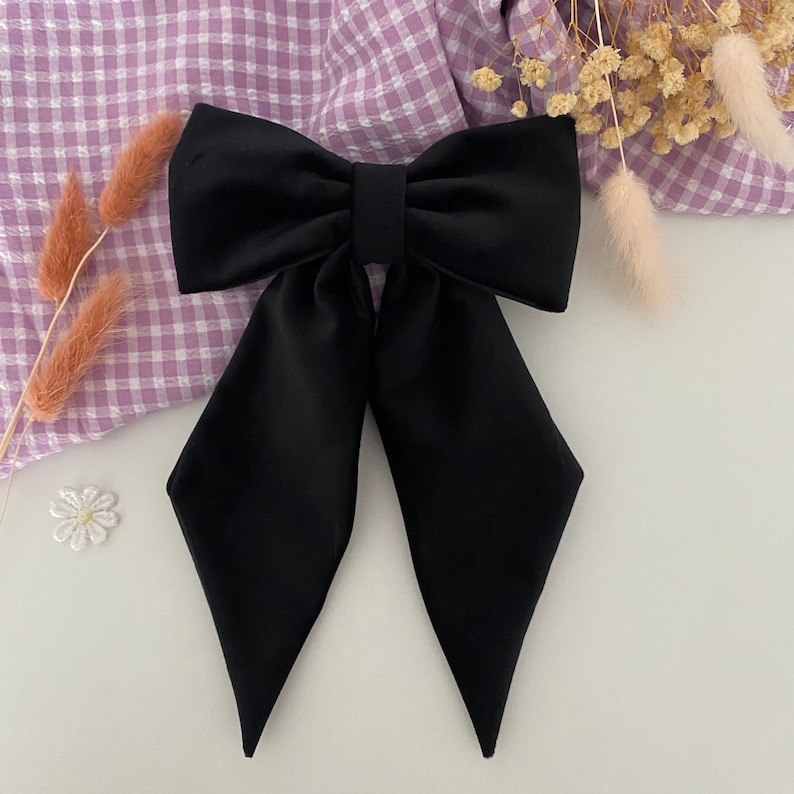 Puede incluir: Un lazo para el cabello de color negro s&oacute;lido con una parte superior ancha y estructurada y dos colas largas y puntiagudas que se extienden hacia abajo. La tela tiene un acabado liso y mate, con un detalle de nudo rectangular en el centro.