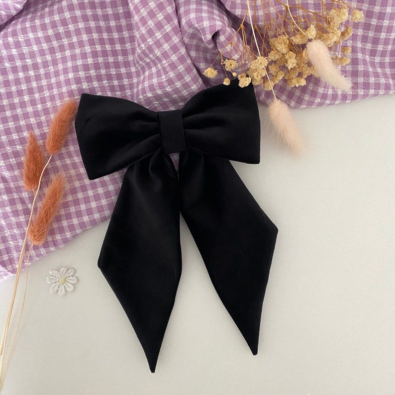 Puede incluir: Un lazo para el cabello de color negro s&oacute;lido que presenta una secci&oacute;n superior ancha y estructurada, junto con dos colas largas y puntiagudas que se extienden hacia abajo. El lazo est&aacute; hecho de una tela lisa y mate con dise&ntilde;o sim&eacute;trico.