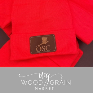 May include: Red knit beanie with a brown leather patch featuring a pheasant silhouette and the text "OSC".
