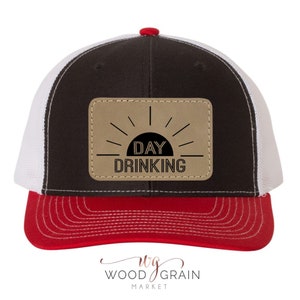 May include: A black, white, and red trucker hat with a brown leather patch that says "DAY DRINKING" with a sun graphic.