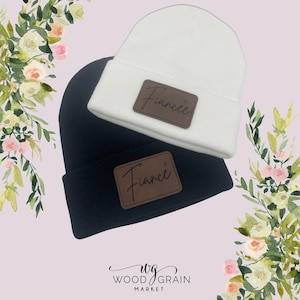 May include: Two beanies, one white and one black, are stacked. Each beanie has a brown leather patch with the word "Fiancée" in script. The beanies are set against a soft pink background with floral accents.