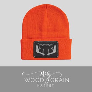 May include: Bright orange beanie hat with a black rectangular patch. The patch features the text "POP-POP" above a silver antler design. The hat is made of a knitted material.