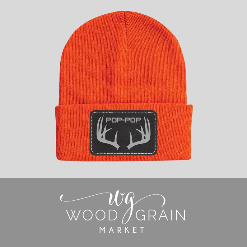 May include: Bright orange beanie hat with a black rectangular patch. The patch features the text "POP-POP" above a silver antler design. The hat is made of a knitted material.