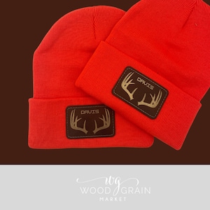 May include: Two bright orange beanies with a brown leather patch featuring the name "DAVIS" and a gold antler design. The beanies are made of a knitted material and are displayed on a brown and white background.
