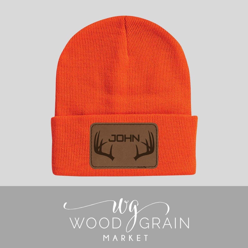May include: Bright orange beanie with a brown leather patch. The patch features the name "JOHN" above a deer antler design. The hat is a warm, winter accessory.