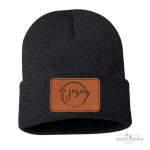 Puede incluir: Gorro negro con un parche de cuero marrón. El parche presenta las palabras "Jesus is King" en un diseño circular con una cruz. El gorro es de un estilo simple y clásico.