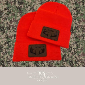 May include: Two bright orange beanies with a brown leather patch featuring the word "CUDDY" above a deer antler design. The beanies are displayed on a camouflage background, showcasing a hunting or outdoor theme.