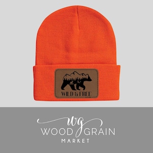 May include: Orange beanie with a brown leather patch featuring a black silhouette of a bear walking in front of mountains with the text "Wild & Free".