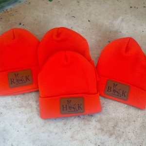May include: Three orange knit beanies with brown leather patches featuring a deer silhouette and the letters "R&K", "B&K", and "H&K".