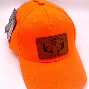 May include: Bright orange baseball cap with a curved brim. A brown leather patch on the front displays the text "LEGENDS LIVE ON" above a deer antler graphic and the numbers "4:29:25". The cap has a structured crown and a button on top.