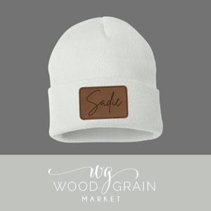 Puede incluir: Un gorro blanco con un parche de cuero marrón. El parche presenta el nombre "Sadie" en escritura cursiva. El gorro está hecho de tejido de punto y se muestra sobre un fondo gris.