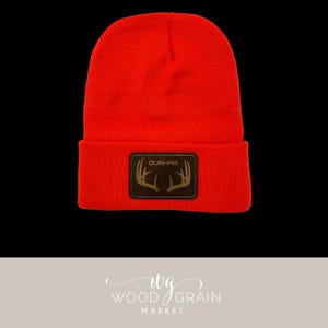 May include: Bright orange beanie with a brown leather patch. The patch features the word "DURHAM" above a deer antler design. The beanie is made of a knitted material.