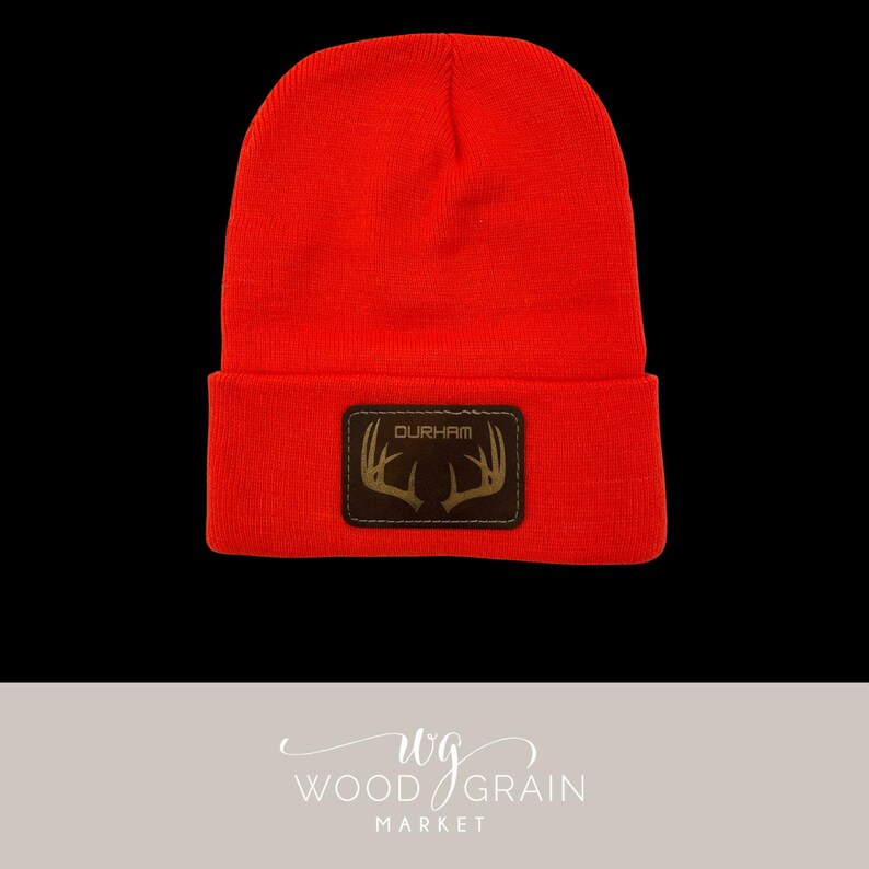 May include: Bright orange beanie with a brown leather patch. The patch features the word "DURHAM" above a deer antler design. The beanie is made of a knitted material.