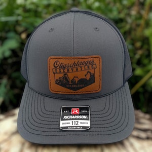 May include: A gray and black trucker hat with a brown leather patch that says "Olischlager Excavating" and a phone number. The hat is on a wooden surface with green foliage in the background.