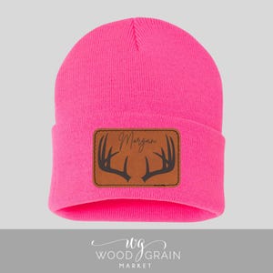 May include: A bright neon pink knit beanie featuring a rectangular brown leather patch on the front cuff. The patch displays a dark brown graphic of deer antlers and the name Morgan in a cursive script font centered above the antlers.