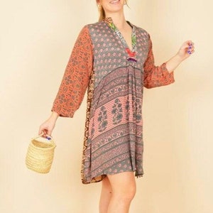 Puede incluir: Una mujer lleva un vestido corto de varios colores con estampado y escote en V. El vestido tiene un diseño de patchwork con diferentes patrones florales y geométricos en tonos rosa, naranja y gris. Lleva una pequeña cesta tejida.
