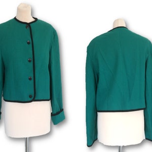 Puede incluir: Una chaqueta corta de color verde azulado con ribetes negros alrededor del cuello, los puños y el dobladillo. La chaqueta tiene cierre de botones.