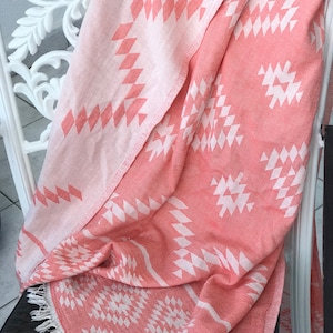 Op de afbeelding: Een roze en witte gestreepte strandhanddoek met een franje rand. De handdoek is gevouwen en over een witte stoel gelegd.