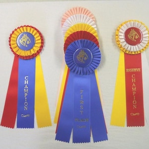 Könnte beinhalten: Eine Sammlung von Pferde-Auszeichnungen in verschiedenen Farben. Die Bänder sind rot, blau, gelb und weiß und tragen die Aufschriften "Champion", "Erster Platz" und "Reserve Champion". Jedes Band hat eine Rosette mit einem Pferd-Reiter-Emblem.
