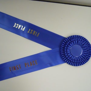Puede incluir: Cinta azul con la inscripción "FIRST PLACE" en letras doradas. La cinta tiene una roseta en el centro. Ideal para ceremonias de premios, competiciones o eventos festivos.
