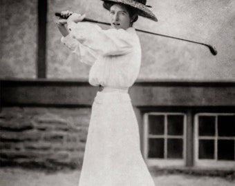 Puede incluir: Fotografía en blanco y negro de una persona con vestido blanco y sombrero, balanceando un palo de golf. La persona está de pie en una zona de césped, con un edificio y ventanas al fondo. El sombrero tiene un ala ancha.
