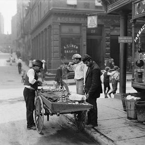 May include: A black and white photo shows a street scene with a vendor selling produce from a cart. The cart is wooden with a metal frame and wheels. It is laden with various fruits and vegetables. Two men stand near the cart, one appears to be the vendor and the other a customer.  A pharmacy is visible in the background with signage in Spanish and English. The street is paved, and there are buildings on either side.  The overall scene depicts a bustling street market in a city.