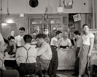 May include: A black and white photo of a group of men sitting at a counter in a diner. The men are wearing casual clothing and some are drinking coffee. There is a sign on the wall that says "We Need America One More Time".