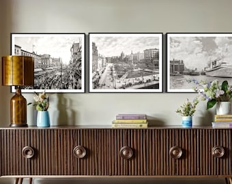 May include: Three black and white framed photographs of cityscapes and a ship on a body of water. The photographs are hanging on a wall above a dark brown wooden cabinet with three drawers.