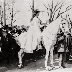 May include: A woman in a white cape rides a white horse through a crowd of people. The woman is wearing a crown and has long hair. The horse is wearing a bridle and has a saddle.