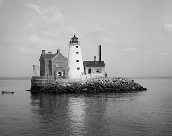 May include: Black and white photograph of a lighthouse on a rocky island. The lighthouse is white with a black top, and there is a stone building next to it. A small boat floats in the water.