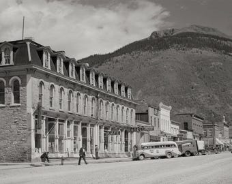 May include: A black and white photo of a vintage hotel in a mountain town. The hotel has a cafe and a sign that says "Cafe Hotel". There are several vintage cars parked in front of the hotel.