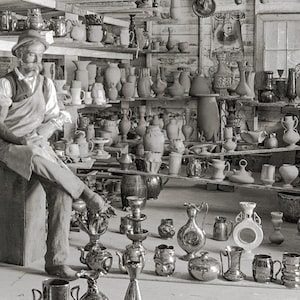 May include: A black and white photo of a pottery workshop with a man sitting on a stool in the foreground. The man is wearing a hat and overalls and is holding a piece of pottery. The workshop is filled with shelves of pottery, including vases, jugs, and bowls.