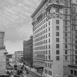 May include: A black and white photograph of a city street with a tall building in the background. The building has many windows and a large entrance. There are cars and people on the street.