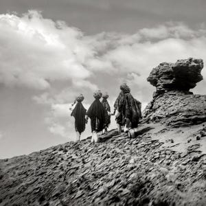 May include: Black and white photograph of four people wearing traditional clothing walking across a rocky landscape towards a large rock formation. The sky is cloudy.