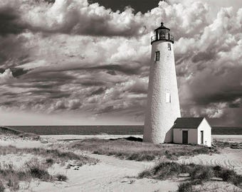 May include: A black and white photograph of a tall white lighthouse with a small white building next to it. The lighthouse is on a sandy beach with tall grass and a cloudy sky.