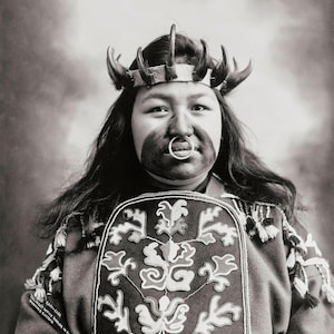 May include: A black and white portrait of a Native American woman wearing a traditional headdress with a beaded design. She is wearing a beaded vest with a floral pattern and has a nose ring.