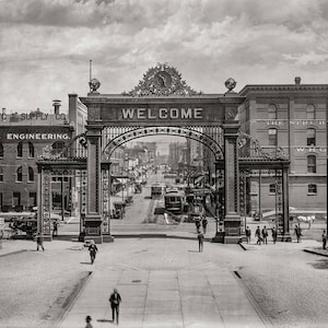May include: A black and white photograph of a street scene in the early 1900s. A large ornate archway with the word "WELCOME" inscribed on it stands in the center of the image. The archway is made of metal and has a decorative design. There are buildings on either side of the archway, and people are walking on the street.