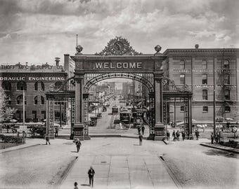 May include: A black and white photograph of a street scene in the early 1900s. A large ornate archway with the word "WELCOME" inscribed on it stands in the center of the image. The archway is made of metal and has a decorative design. There are buildings on either side of the archway, and people are walking on the street.