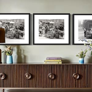 May include: Three black framed monochrome prints depicting vintage street scenes.  The central image shows a bustling marketplace with vendors and shoppers, while the flanking images showcase similar scenes.  The prints are displayed on a dark brown ribbed wood media console with brass hardware.  The console also features a gold table lamp, blue vase, and books.  The overall aesthetic is mid-century modern.
