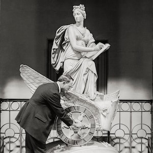May include: A man in a suit adjusts the hands of a clock on a winged chariot sculpture. The sculpture is of a woman holding a book, and is made of white marble.