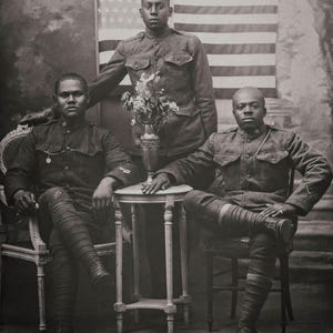 May include: Three men in military uniforms pose for a portrait. They are seated in front of an American flag. The men are wearing hats and have serious expressions on their faces.