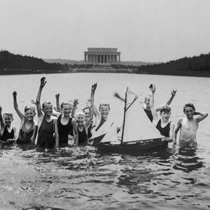 May include: A group of children play in a lake with a small sailboat. They are all wearing swimsuits and are smiling. The Lincoln Memorial is in the background.