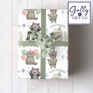 May include: Gift wrap featuring a repeating pattern of cartoon cats with party hats, balloons, and the words "Happy Birthday." The wrapping paper is tied with a sage green ribbon and bow. The Golly Gift Co. logo is in the upper right corner.