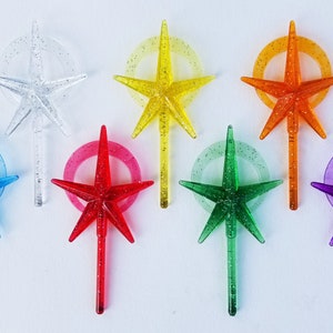 May include: A collection of eight star-shaped ornaments in various colors, including clear, yellow, orange, red, green, blue, and purple. Each star has a glittery finish and is attached to a stem with a circular accent. These ornaments are ideal for holiday decorations.