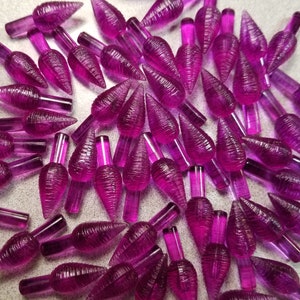 May include: A collection of translucent, purple Christmas light bulbs. Each bulb has a conical shape with vertical ridges and a cylindrical base. The bulbs are scattered across a light-colored surface.