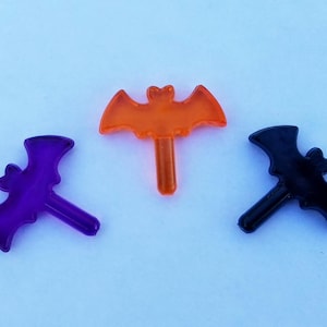 May include: Three plastic Halloween decorations shaped like bats. One is orange, one is purple, and one is black.