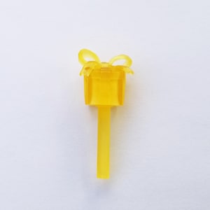 May include: A translucent yellow gift box-shaped toothpick with a bow on top. The toothpick has a rectangular box design with a bow on top and a long, thin stem. The background is plain white.