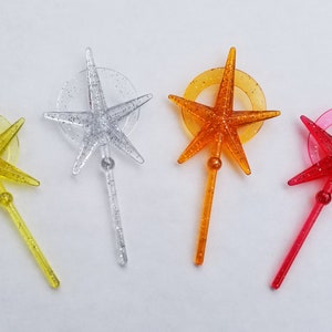 May include: Four colorful star-shaped cocktail picks. Each pick features a star with glitter accents, a circular ring, and a stem. Colors include yellow, silver, orange, and red. These decorative picks are ideal for themed parties.