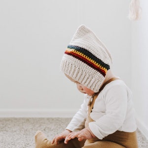 Puede incluir: Un bebé con un gorro de crochet blanco con una franja de arcoíris y polainas marrones. El bebé está sentado en una alfombra.