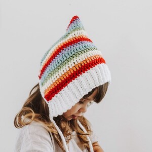 Puede incluir: Un niño lleva un gorro de punto blanco y de rayas arcoíris con lazos largos. El gorro tiene una punta puntiaguda y está hecho con un hilo suave y texturizado.